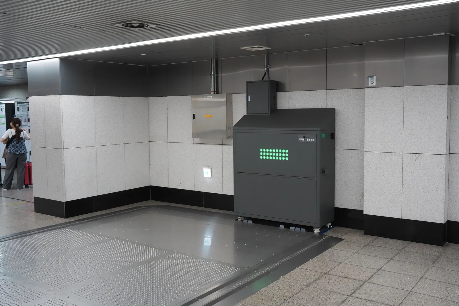 A grey cabinet in a Seoul subway concourse with a grid of green status lights, beside a large metal floor plate set flush into the ground.