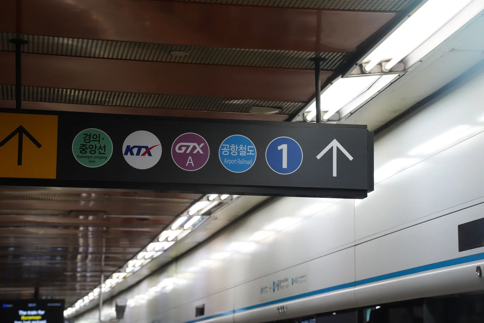 A black sign hanging from a station ceiling showing colored circular markers for the Gyeongui-Jungang Line, KTX, GTX-A, Airport Railroad and Line 1, with an arrow.