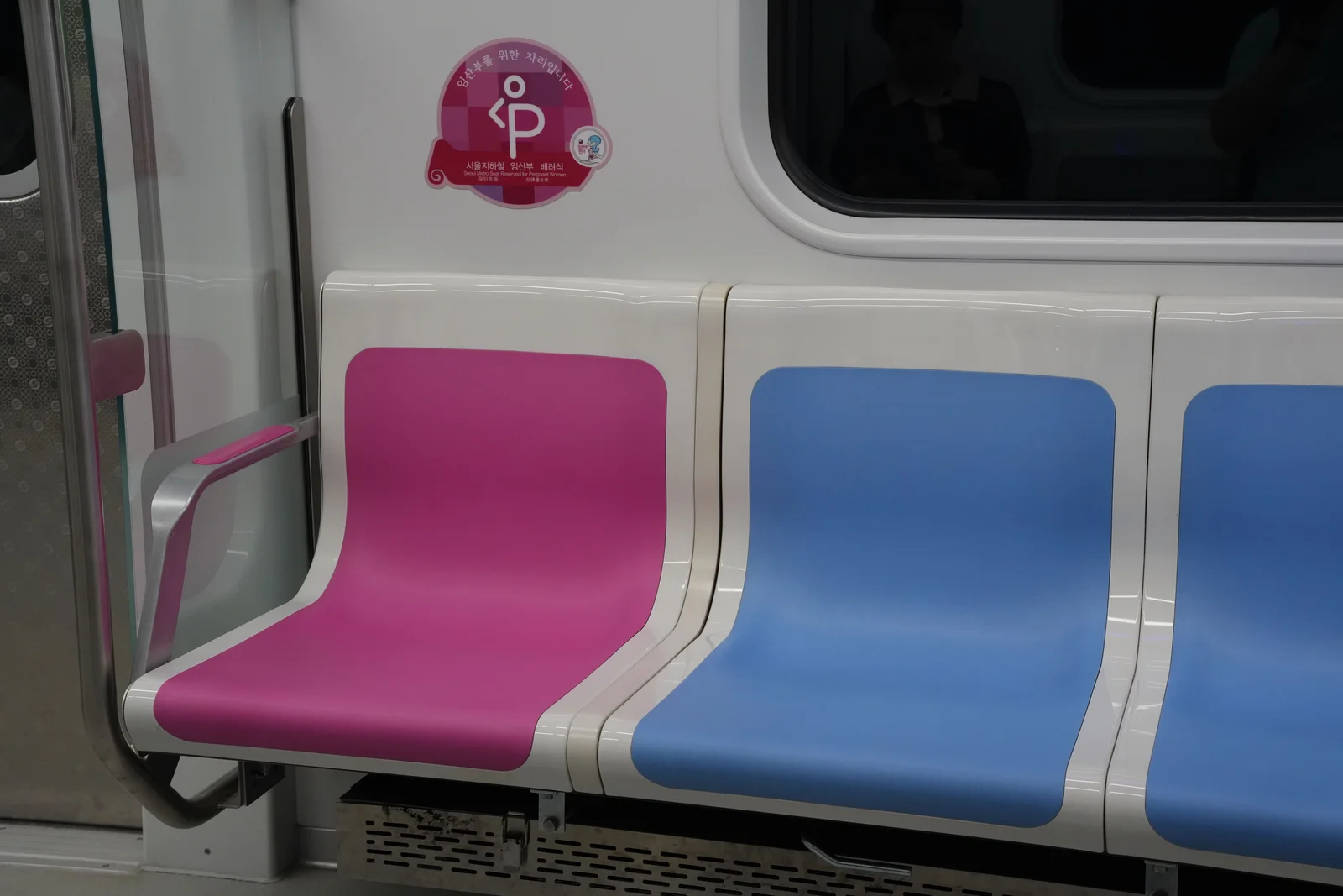 A subway car interior with a single bright pink seat beside blue seats, and a pink decal above it reading Seoul Metro Seat Reserved for Pregnant Women.